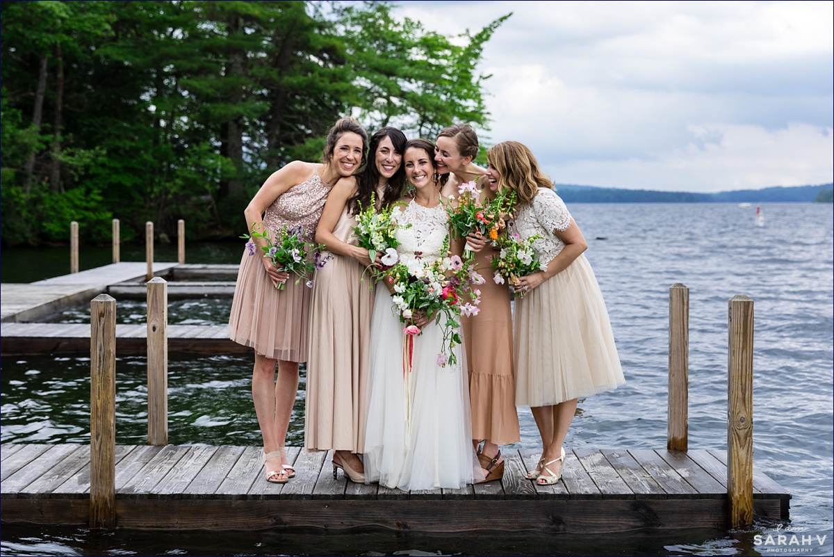 Church Island wedding Ceremony the bride and her bridesmaids in their various soft blush colored dresses embrace on one of the docks on Squam Lake NH