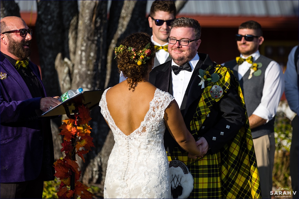 The groom in his kilt smiles warmly at his bride during the outdoor Maine wedding ceremony