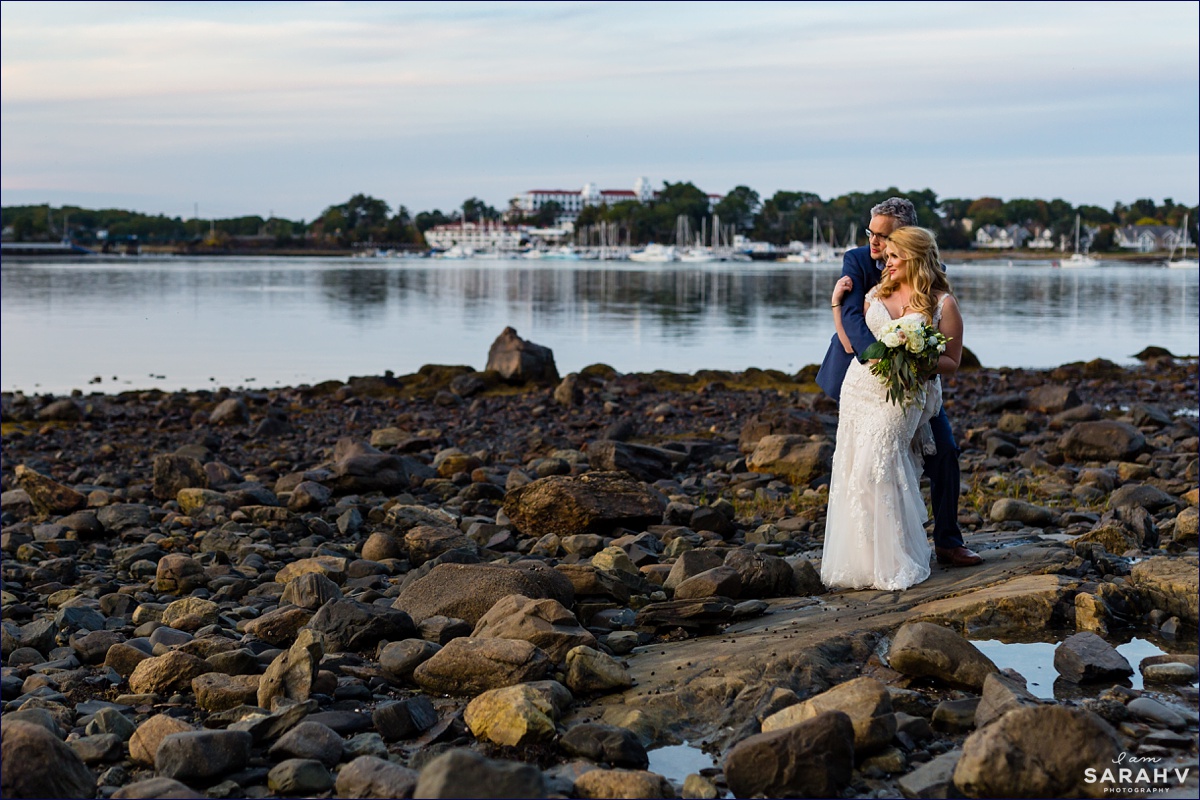 New Hampshire Elopement Photographer the newlyweds stand on the rocky shore with  Wentworth by the Sea in the background