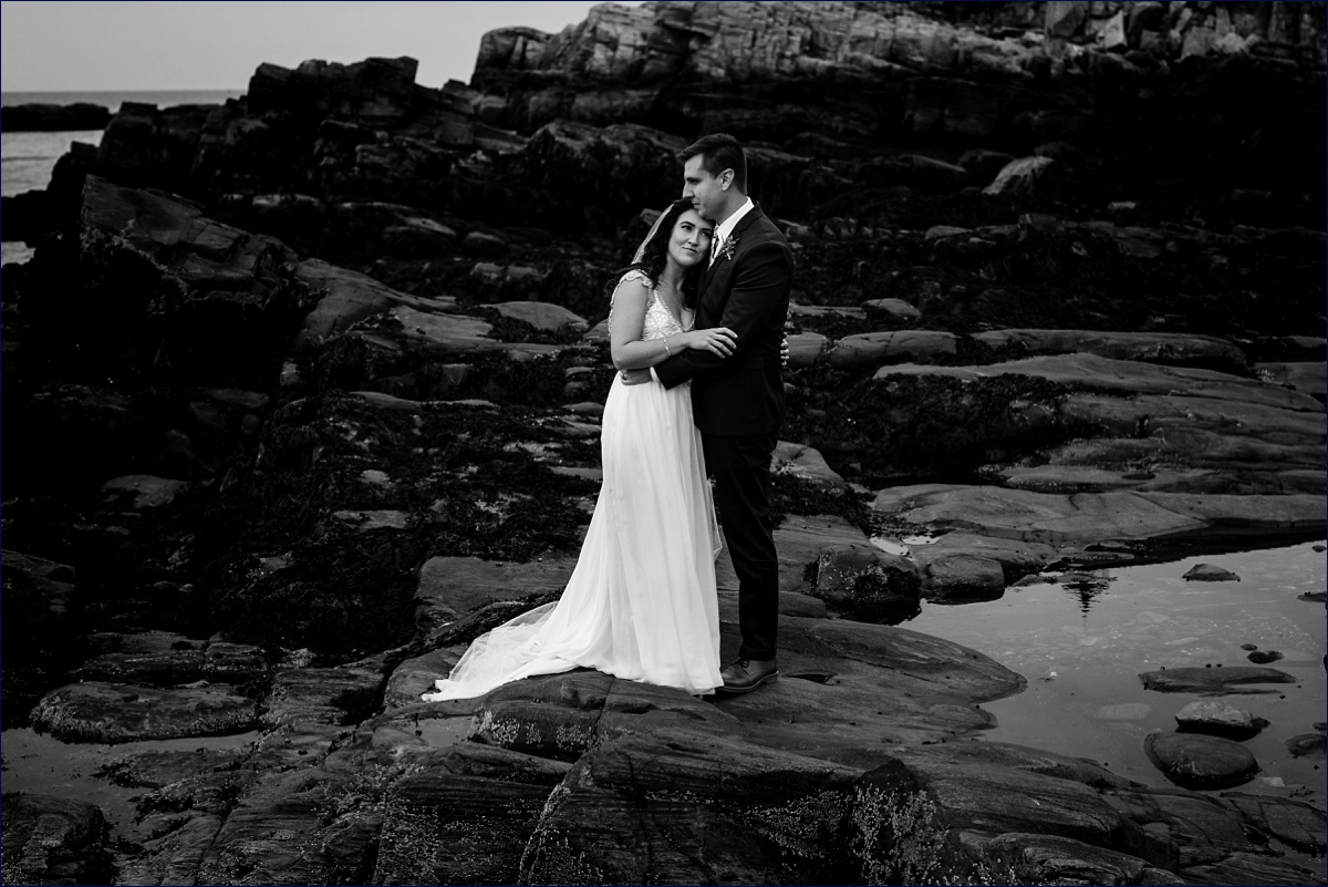 The newly married couple hold each other peacefully against the dark rocks of Cape Elizabeth Maine on their elopement day