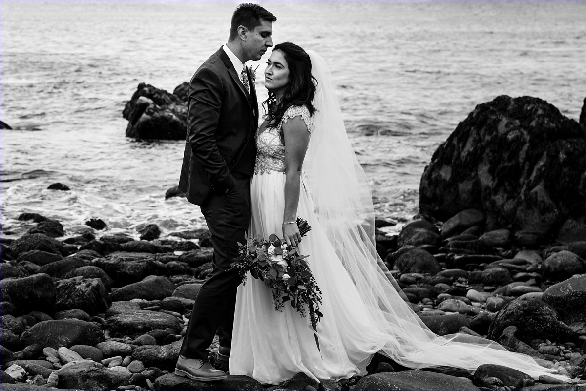 The groom kisses the bride on the side of the face as they stand at the water's edge in Portland Maine