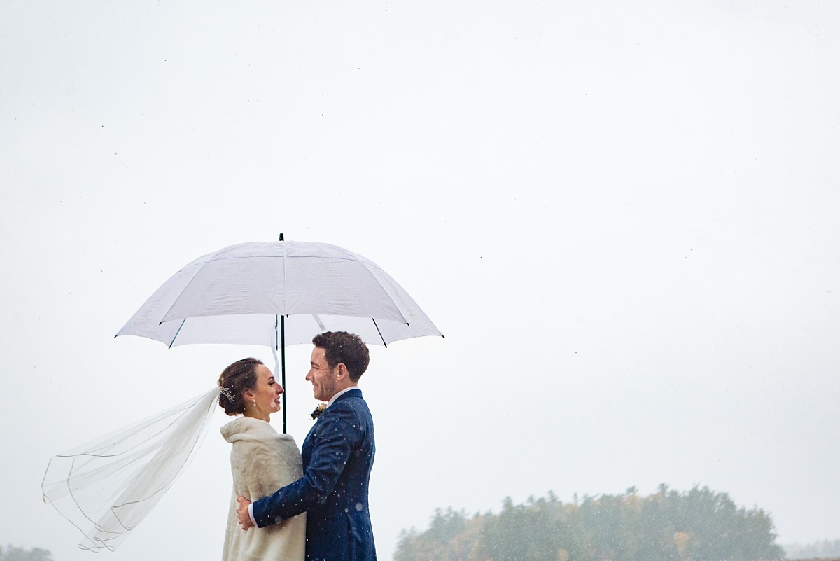 The bride and groom stay under an umbrella in the snow at Migis Lodge on their Maine wedding day