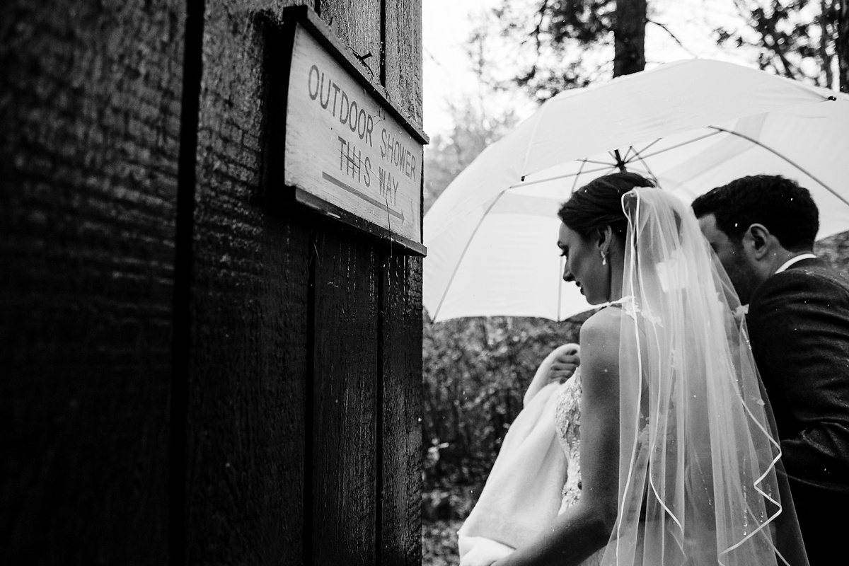 The bride and groom huddle under an umbrella and walk past a sign for an outdoor shower at Migis Lodge in Maine