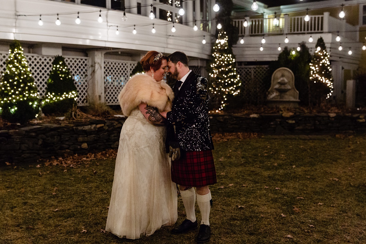 The bride and groom stand close together in the chilly December night after their intimate wedding with his sequin jacket glittering under the bistro lights in New Hampshire