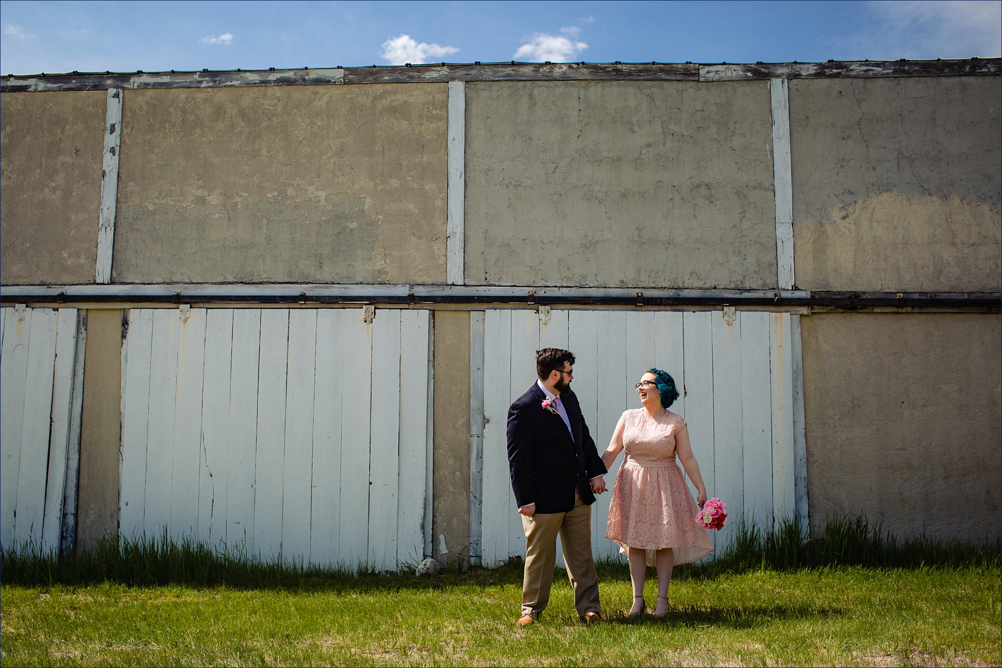 The groom and bride share a laugh in front of a garage at his family's Maine farmhouse