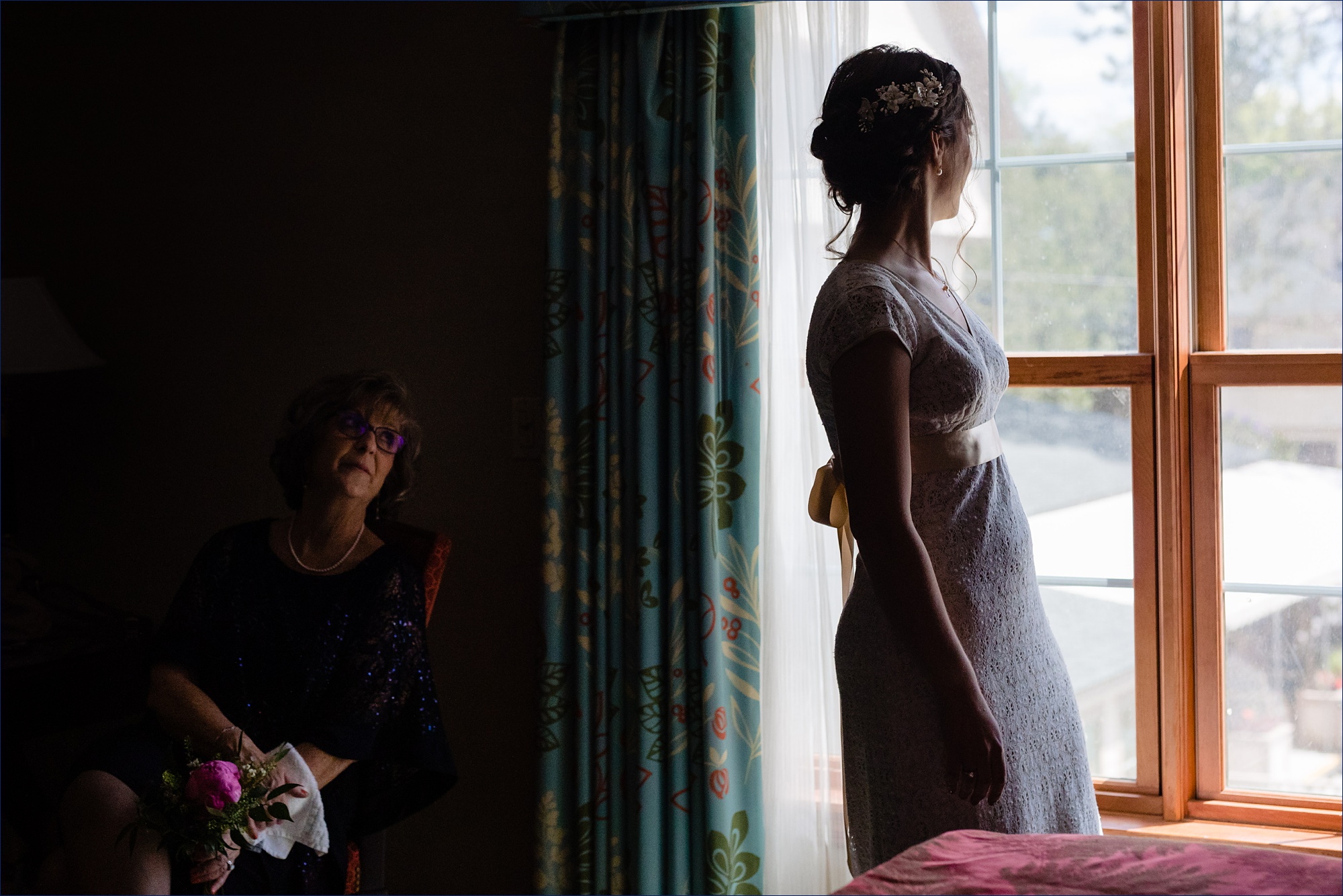 The bride looks out the window as her mom watches her 