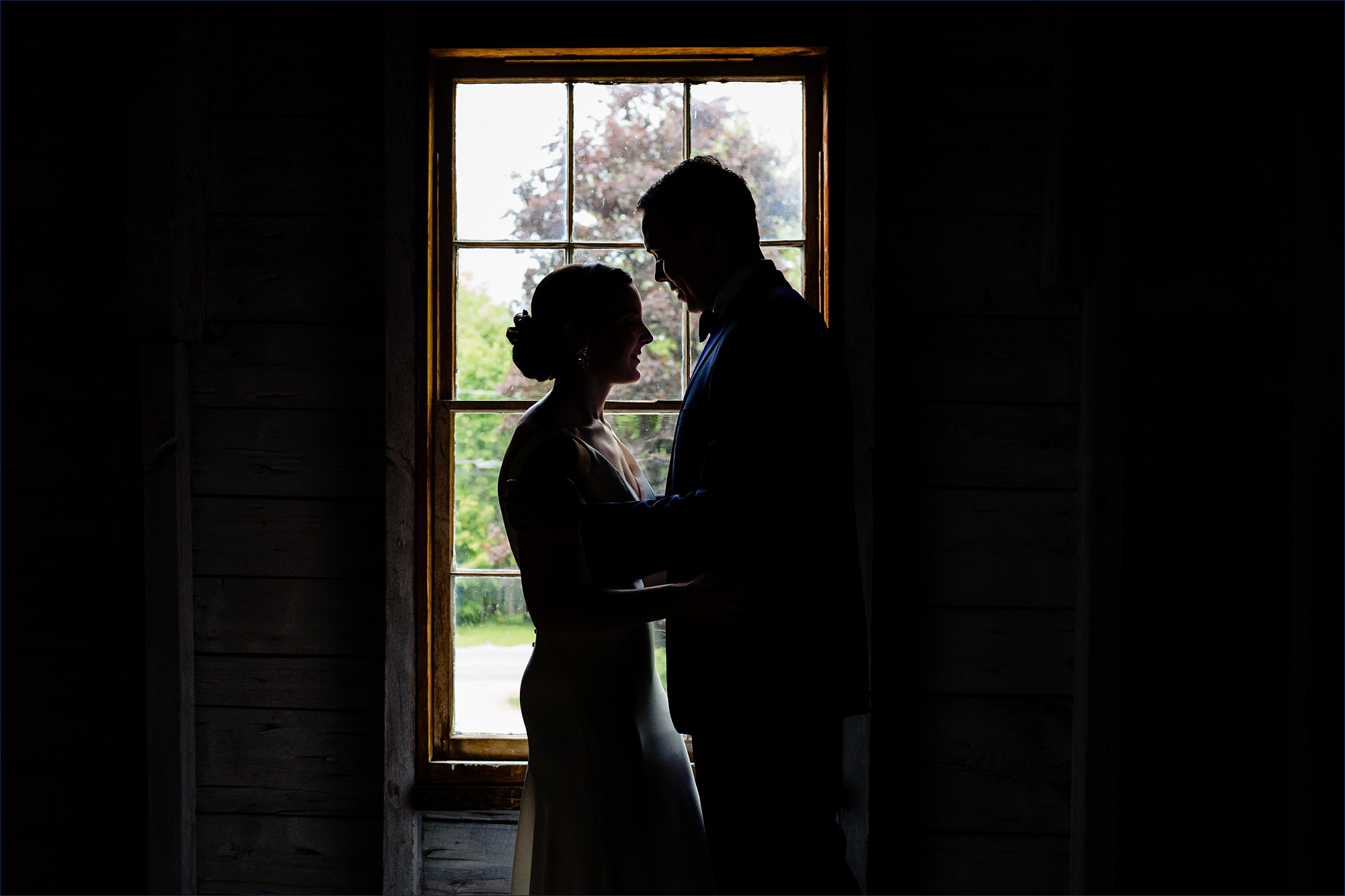 The bride and groom are silhouetted against the barn window in the loft of Maine