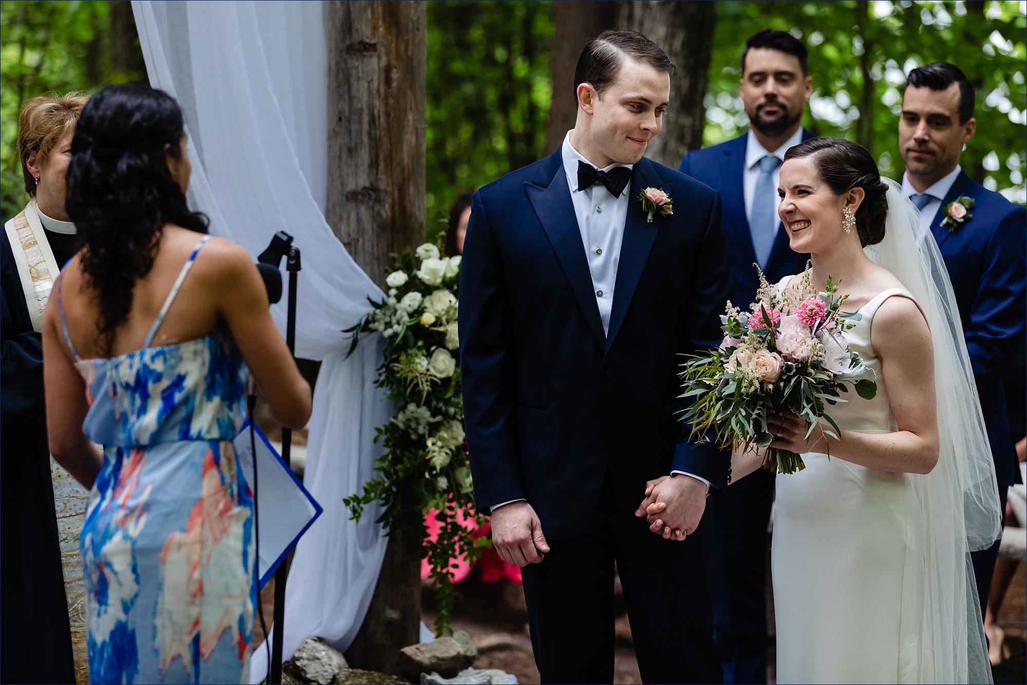 The groom smiles at the bride as they listen to a reading at their outdoor wooded wedding in Maine
