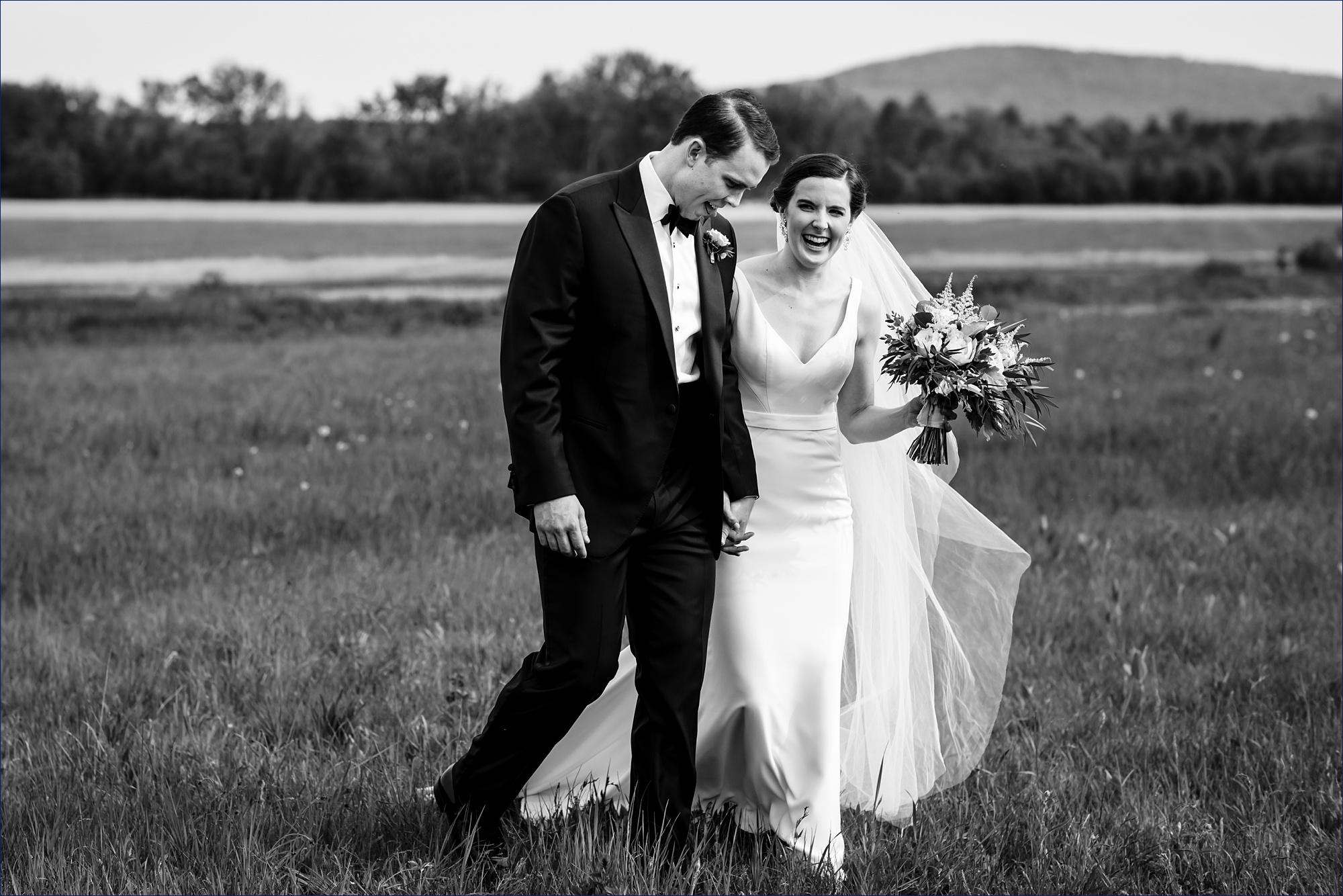 The bride and groom laugh as they walk through the tall grass at the Maine wedding