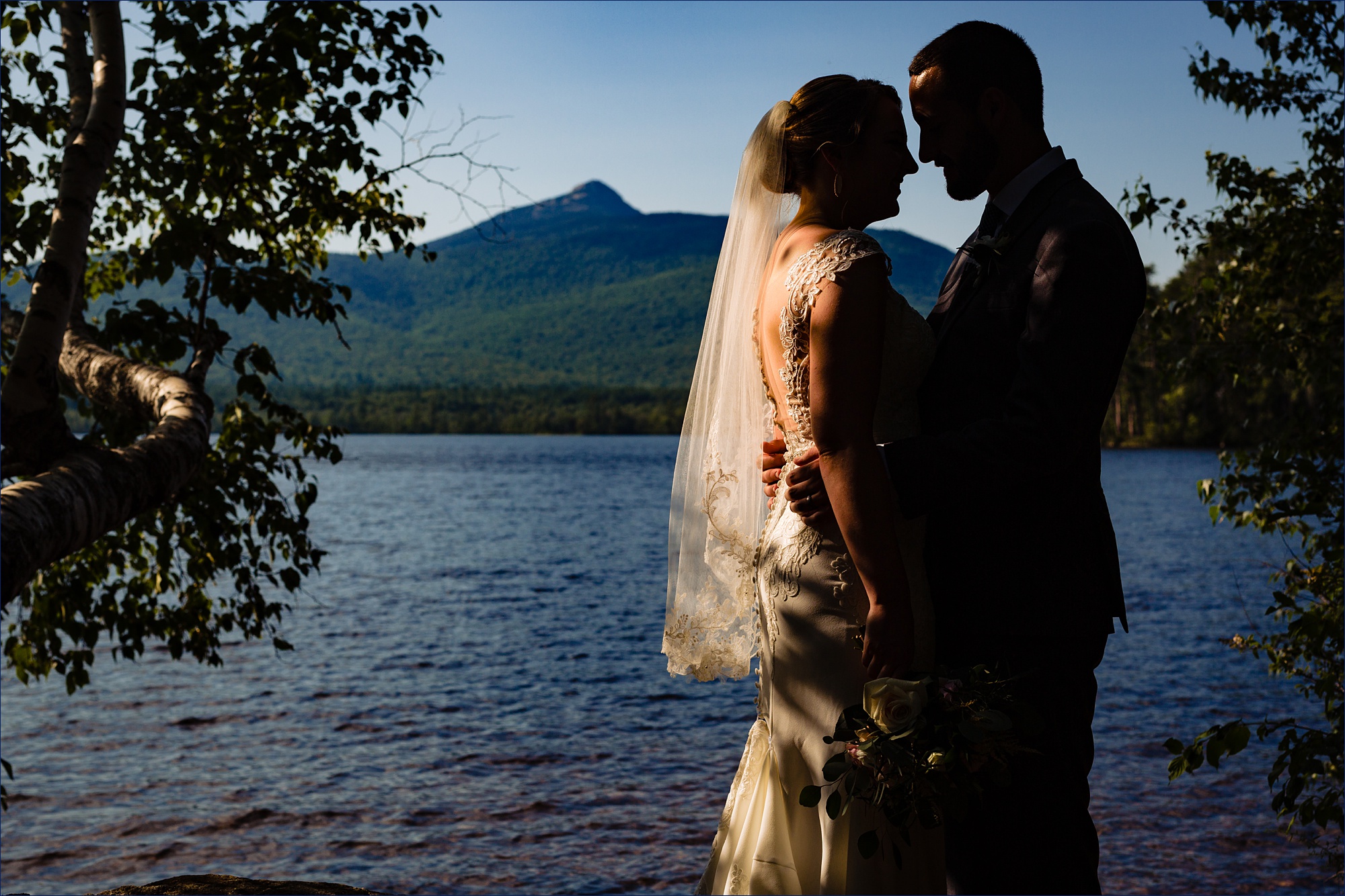 The bride and groom are silhouetted against a bright summer sky with Mount Chocorua standing tall in the distance on their NH wedding day