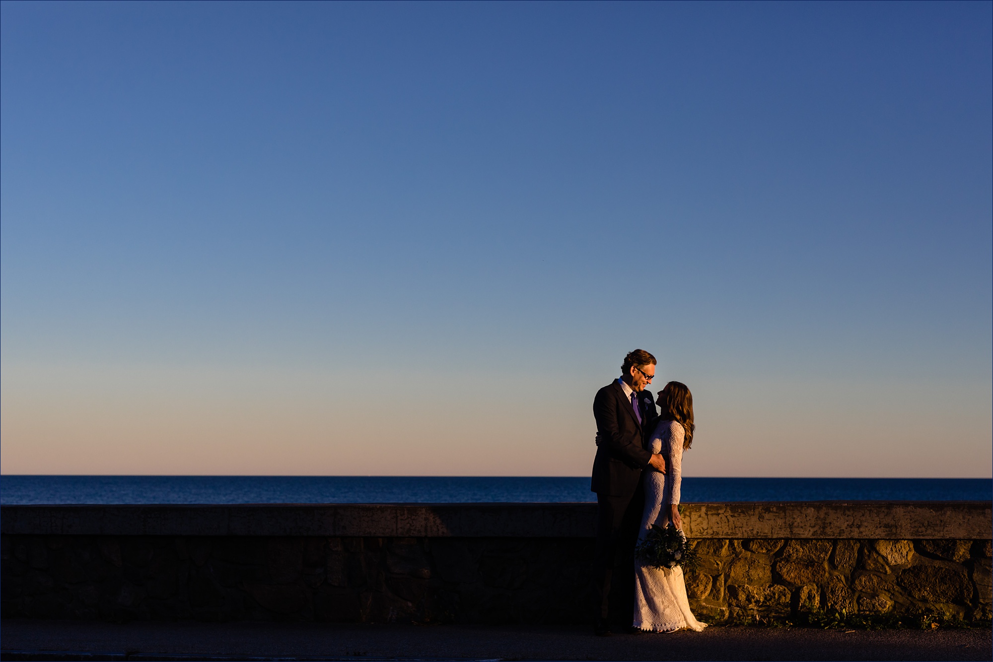 The bride and groom stand in front of the ocean as the sun sets behind them in Kennebunkport Maine