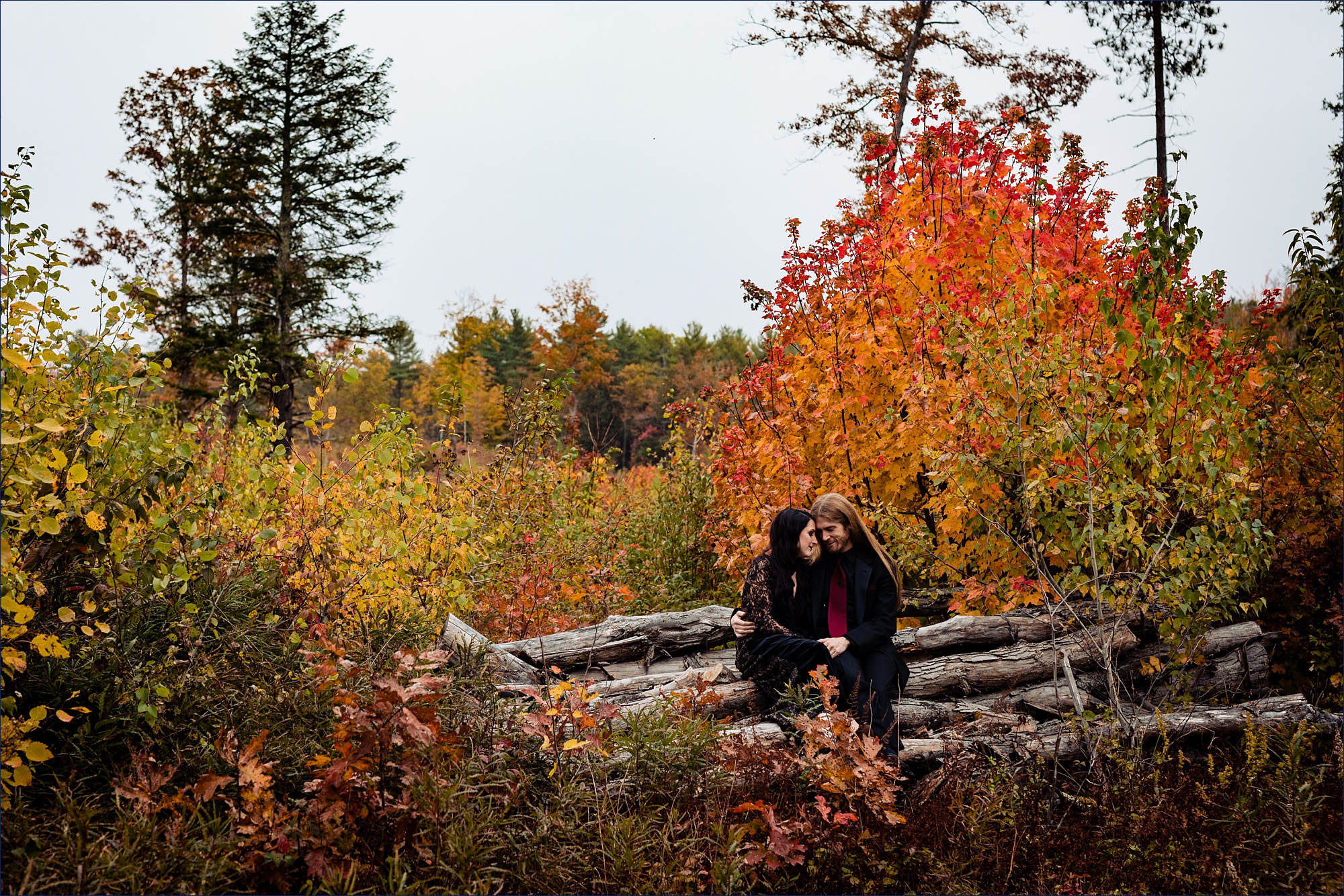 The bride and groom sit among the fall colored leaves and get close together on their New Hampshire wedding day