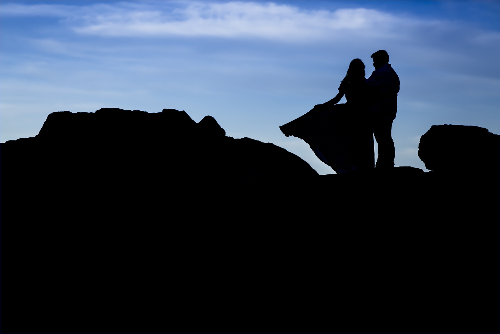 The couple stand on the rocks with a brilliant blue sky at The Cliff House in Maine