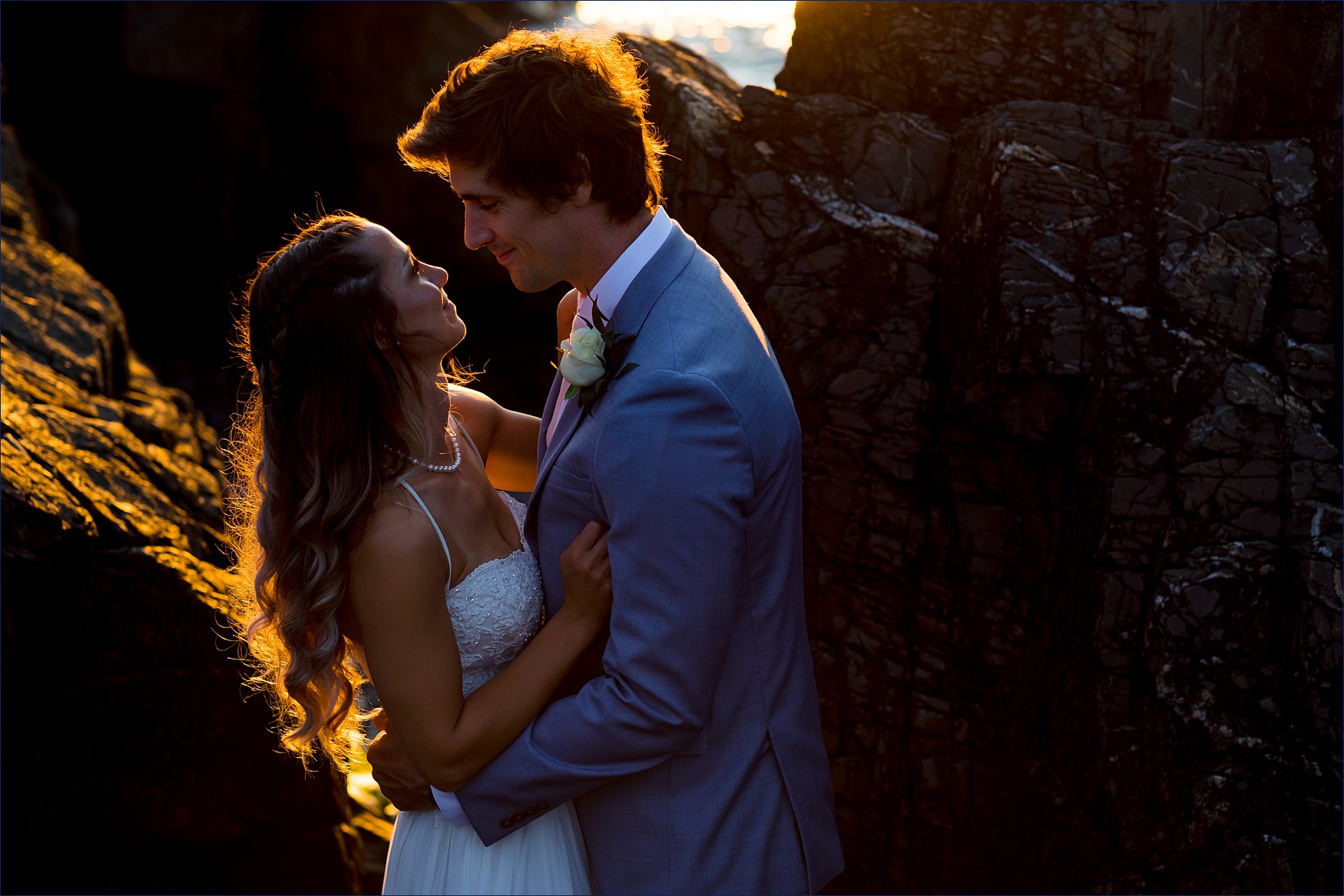 After a Maine elopement, the couple stands close together out in the rocks while the sunrise bathes them in light 