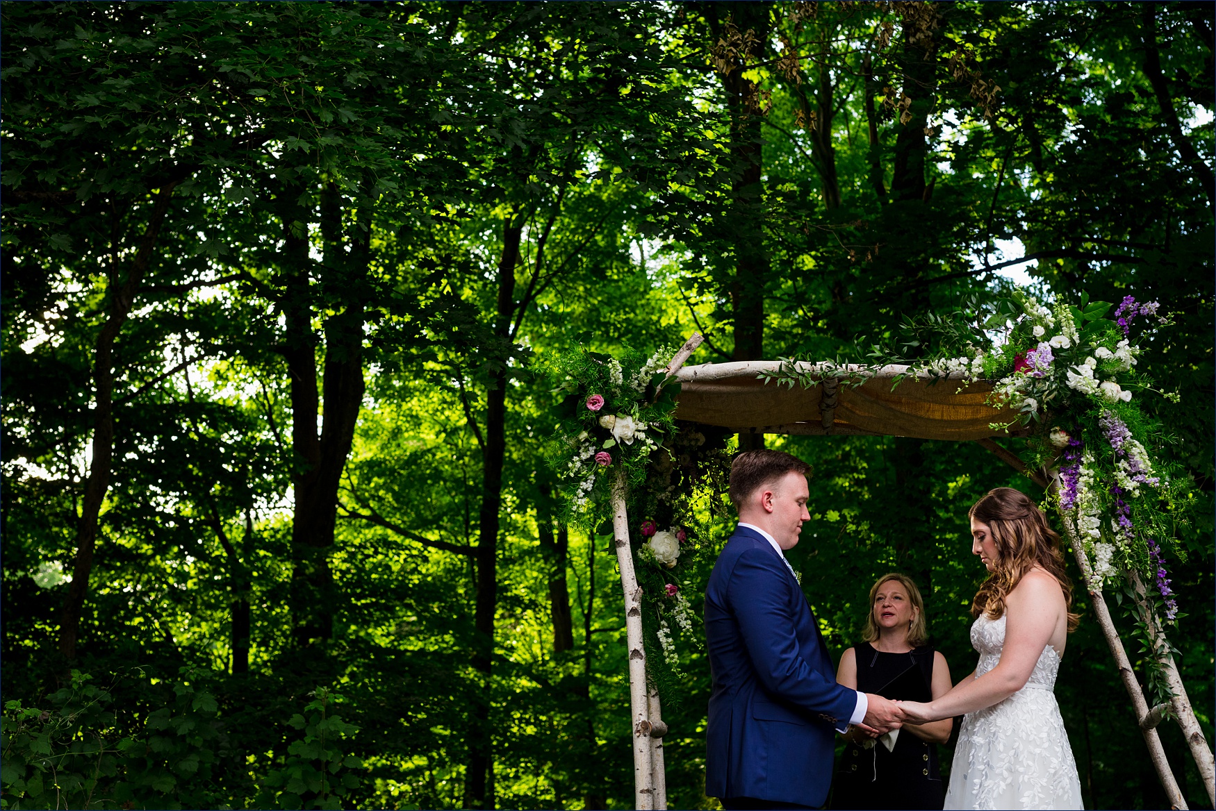 The bride and groom stand among the tall trees on the wedding day