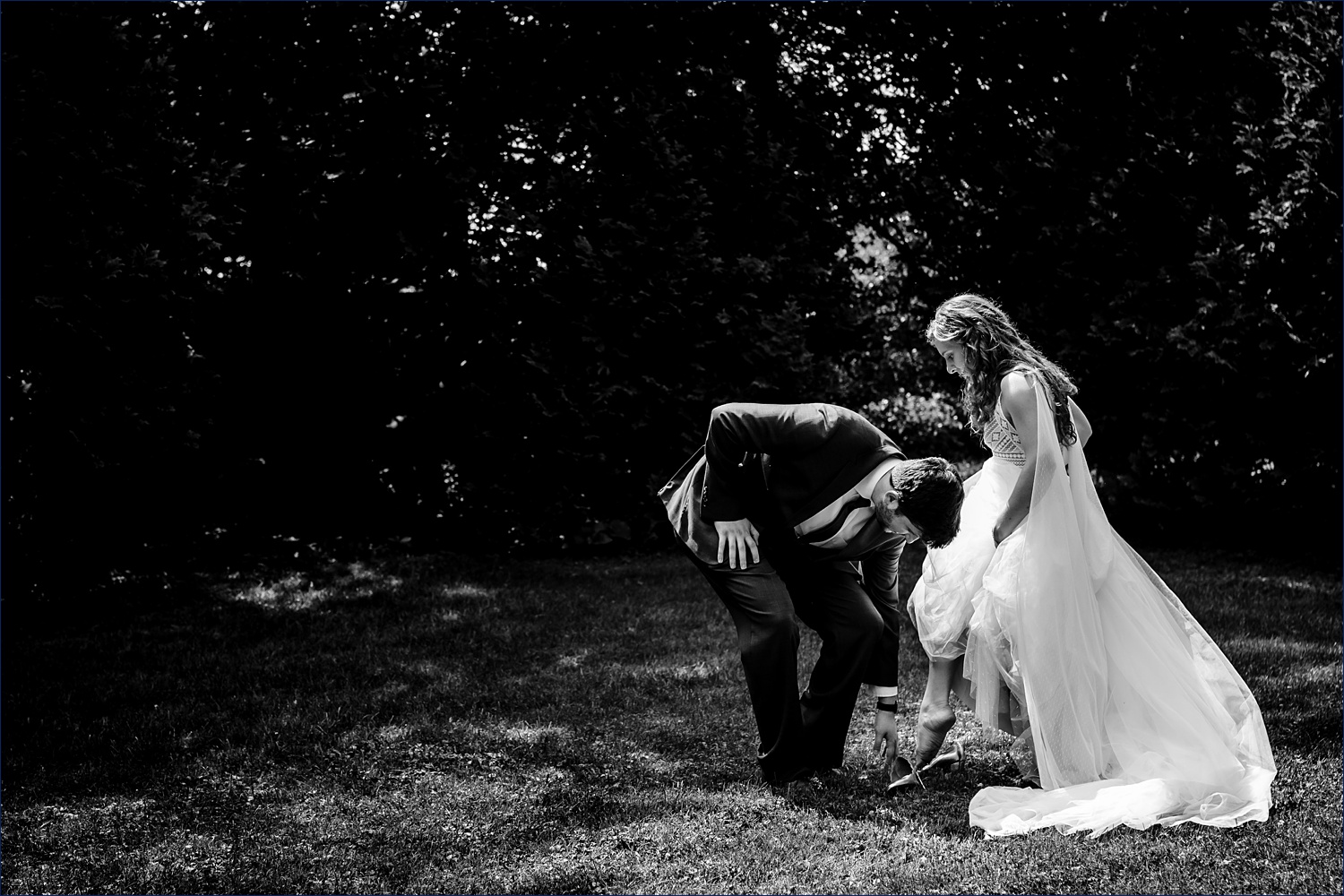 The groom helps the bride into her wedding day shoes out in the grass