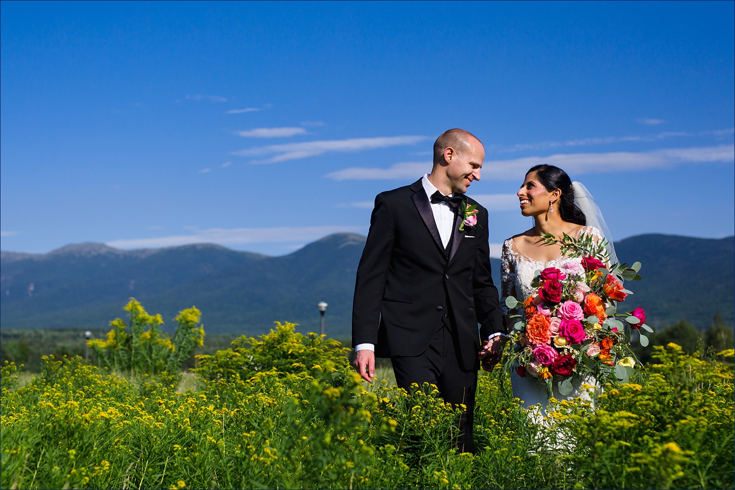 The newlyweds walk out in a meadow with the White Mountains behind them in NH