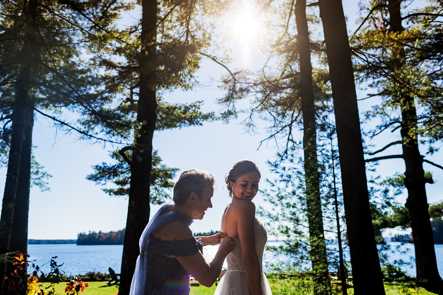 The bride and her mom get her into her wedding dress at Migis Lodge Resort in Maine with Sebago Lake in the distance