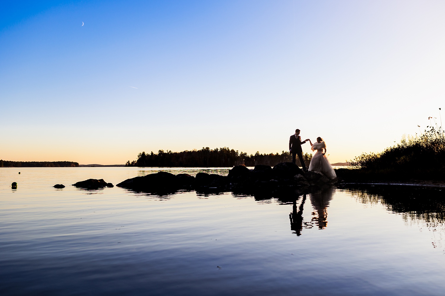 The couple is silhouetted against the setting sun and Sebago Lake on their Maine wedding day 