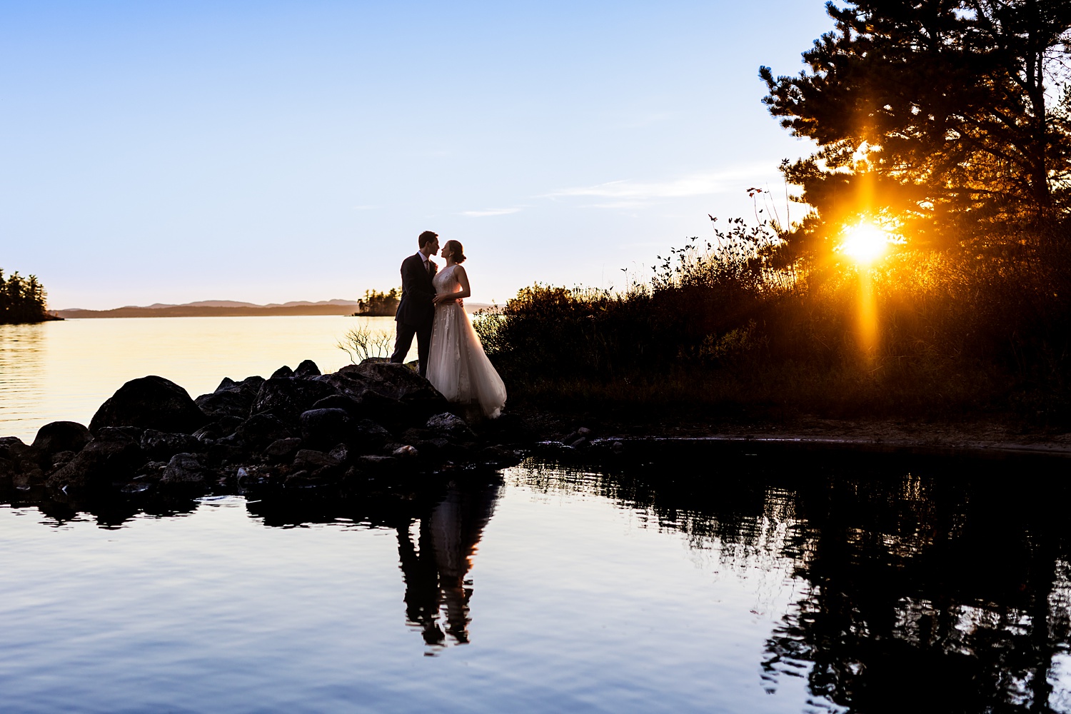 The couple is silhouetted against the setting sun and Sebago Lake on their Maine wedding day 