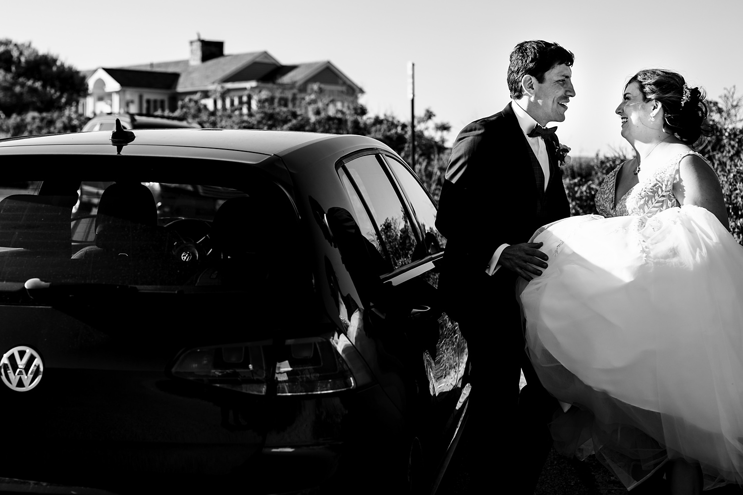 Bride and groom smile as he helps her get into the car on their wedding day