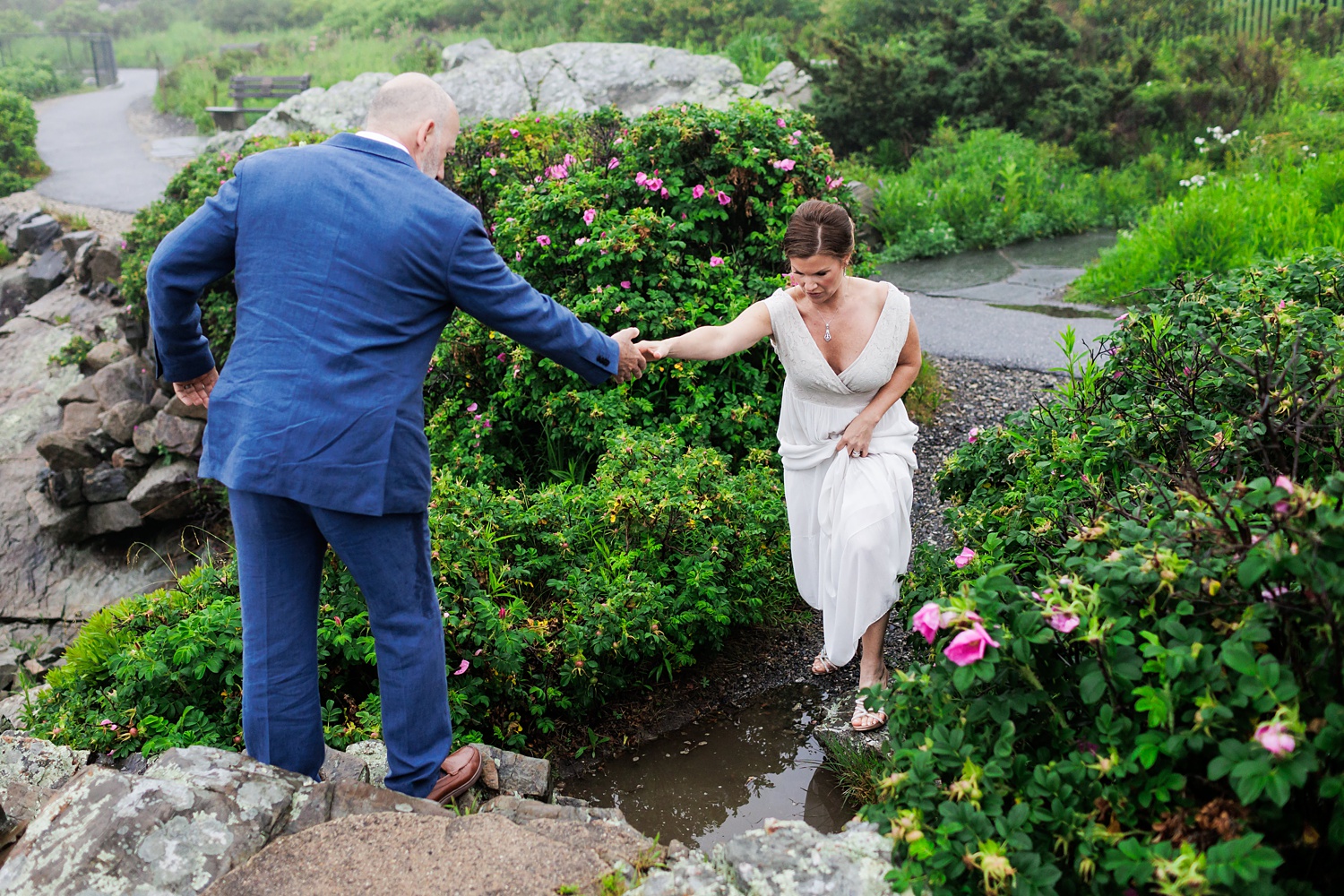 The groom helps the bride over a puddle on their rainy Marginal Way Maine elopement